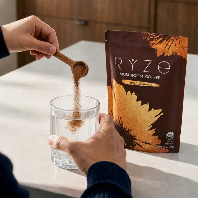 Pouring RYZE mushroom coffee powder into a glass of water beside the pouch on a kitchen counter
