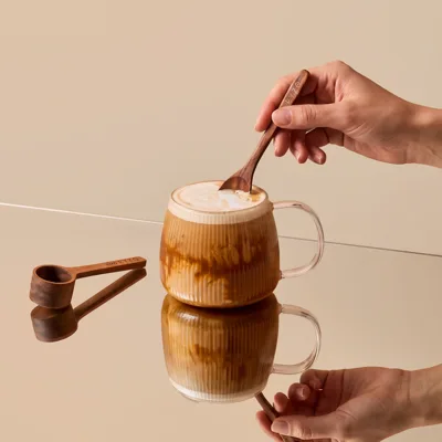 Hand stirring RYZE mushroom coffee into a glass mug with a wooden scoop alongside