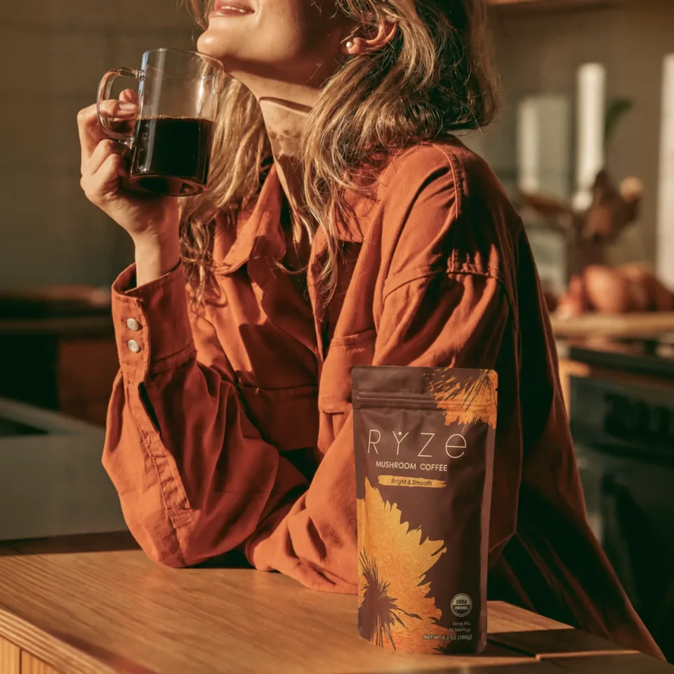 Woman sipping RYZE mushroom coffee from a glass mug beside the bag