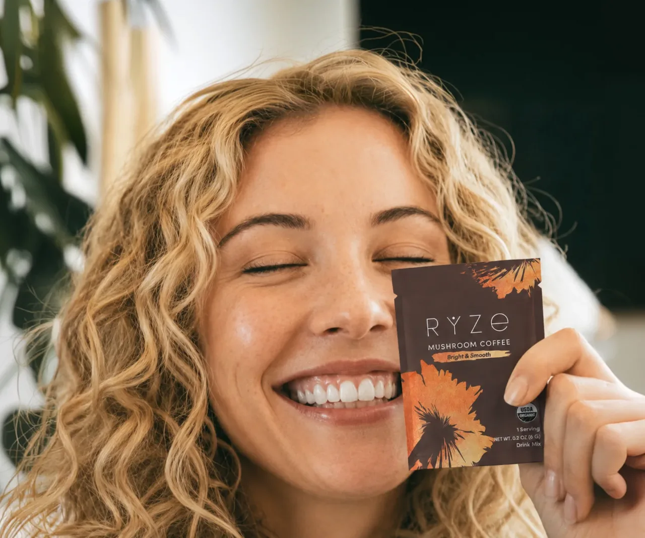 Smiling woman holding a RYZE Mushroom Coffee sachet up to her cheek