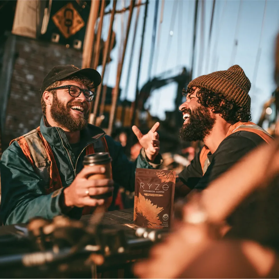 Two crew members laughing over coffee on site, a bag of RYZE Mushroom Coffee on the table between them