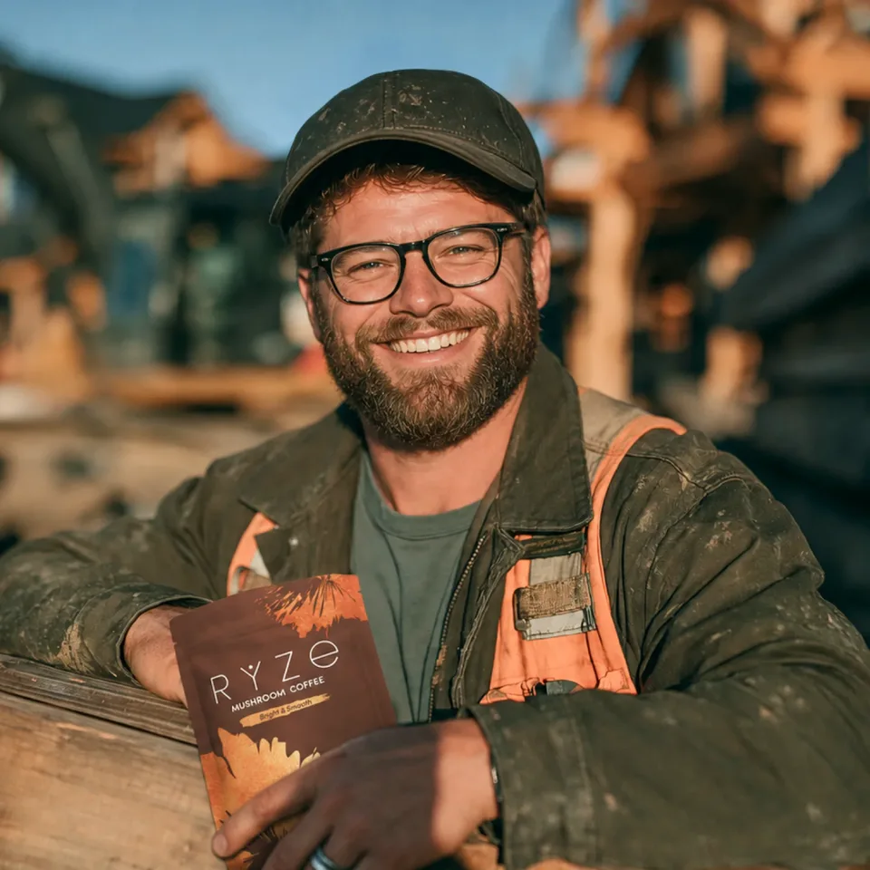 Smiling tradesman in a work cap and hi-vis vest at a framing site, holding a bag of RYZE Mushroom Coffee