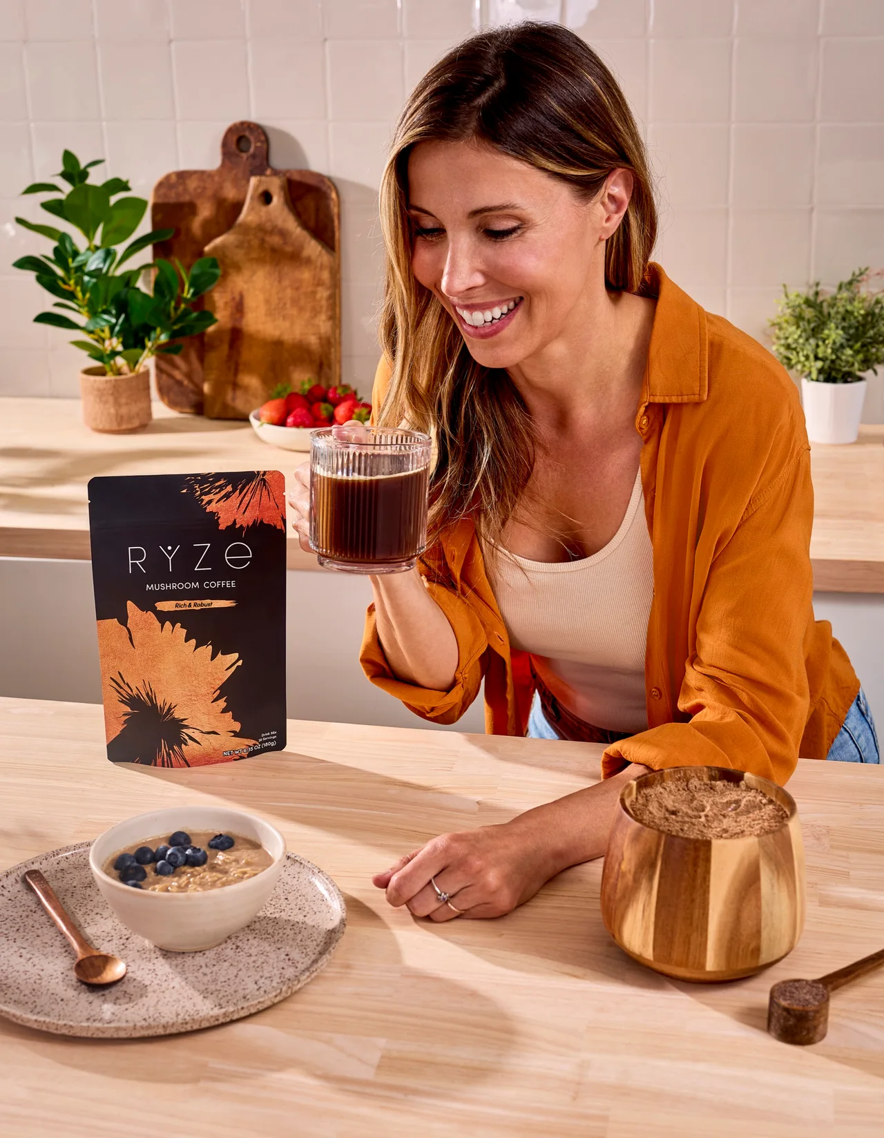 Woman smiling in her kitchen beside a bag of RYZE mushroom coffee and an iced brew