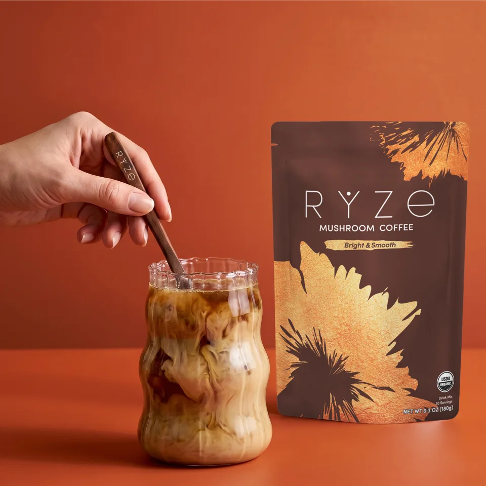 A hand stirring a glass of iced RYZE Mushroom Coffee beside the pouch
